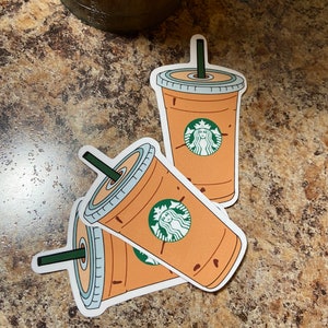 May include: Three stickers featuring a Starbucks iced coffee cup design. The cup is light brown with a green straw and the Starbucks logo. The stickers are on a brown speckled surface.