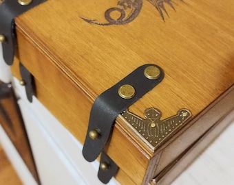 May include: A rectangular wooden box with a light brown finish featuring a dark laser-engraved dragon design on the lid. The box is accented with black leather straps secured by brass-coloured metal studs and decorative brass corner protectors.