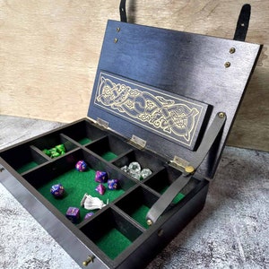Könnte beinhalten: Eine schwarze Holzkiste mit einem Scharnierdeckel und Lederriemen. Die Kiste hat ein grünes Filz-Innenleben mit Fächern für Würfel und anderes Spielzubehör. Der Deckel zeigt ein goldfarbenes keltisches Knotendesign.