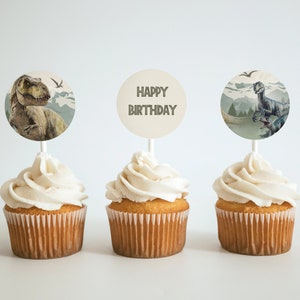 May include: Three cupcakes with white frosting topped with round dinosaur-themed cupcake toppers. The toppers feature a green dinosaur, a blue dinosaur, and the text "Happy Birthday".