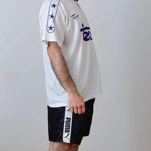 Op de afbeelding: Een wit poloshirt met korte mouwen, een paarse kraag, paarse sterren op de mouwen en de tekst "Tagy" en "Tex" in het paars op de borst gedrukt. Gecombineerd met een zwarte sportshort met een witte zijstreep en het Puma-logo.