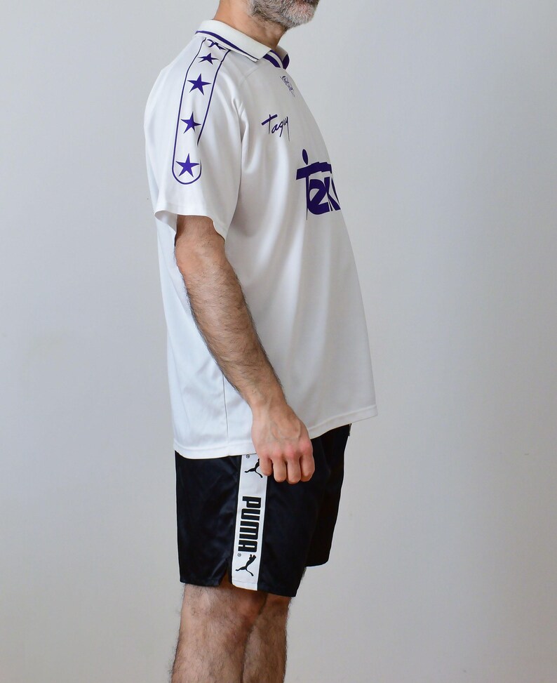Op de afbeelding: Een wit poloshirt met korte mouwen, een paarse kraag, paarse sterren op de mouwen en de tekst "Tagy" en "Tex" in het paars op de borst gedrukt. Gecombineerd met een zwarte sportshort met een witte zijstreep en het Puma-logo.