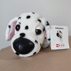 May include: A plush Dalmatian dog toy with white fur and black spots, featuring large dark eyes and a prominent black nose. The toy has pink-lined ears and comes with a white tag that reads Dalmatian, The Dog, and Artist Collection.