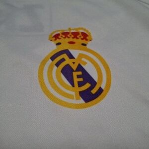 Op de afbeelding: Een wit kledingstuk van stof met het embleem van voetbalclub Real Madrid. Het logo toont een gouden kroon bovenop een gouden cirkel met een diagonale paarse streep en de in elkaar grijpende letters MCF in goud op de witte ondergrond.