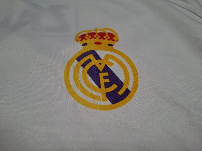 Op de afbeelding: Een wit kledingstuk van stof met het embleem van voetbalclub Real Madrid. Het logo toont een gouden kroon bovenop een gouden cirkel met een diagonale paarse streep en de in elkaar grijpende letters MCF in goud op de witte ondergrond.