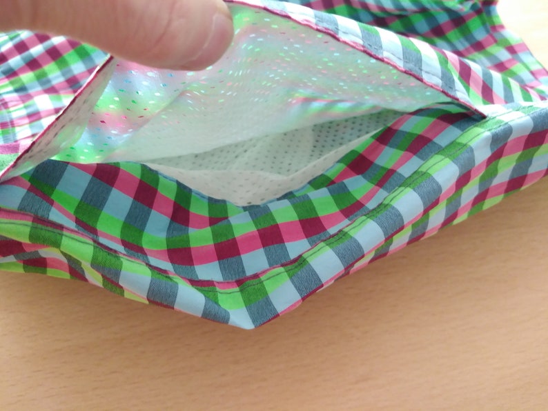 May include: A close-up of a green, pink, and blue plaid fabric with a white mesh pocket lining. The pocket is open and the fabric is folded over.