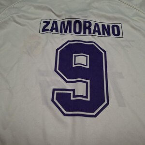 Op de afbeelding: Een wit sportshirt met de naam ZAMORANO in donkerpaarse hoofdletters boven een groot donkerpaars nummer 9 met een witte omlijning. De stof is glad en lichtgewicht en toont een strak atletisch ontwerp op de achterzijde.