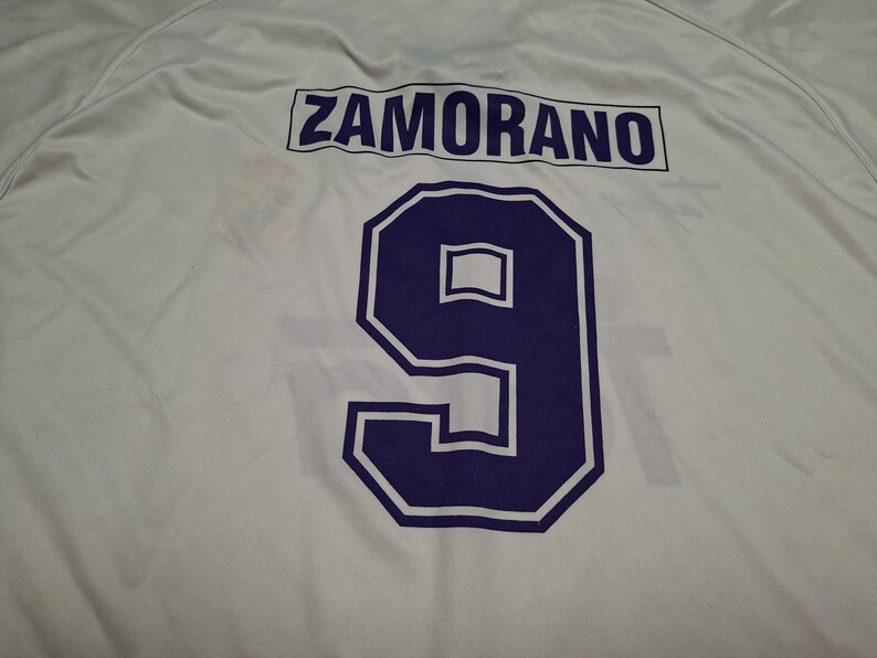Op de afbeelding: Een wit sportshirt met de naam ZAMORANO in donkerpaarse hoofdletters boven een groot donkerpaars nummer 9 met een witte omlijning. De stof is glad en lichtgewicht en toont een strak atletisch ontwerp op de achterzijde.