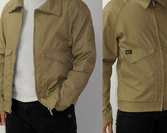 Può includere: Una giacca color beige con chiusura a zip e una tasca a toppa sul lato sinistro. La giacca ha un colletto e maniche lunghe.
