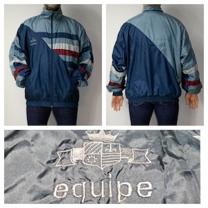 Könnte beinhalten: Eine blaue und graue Windbreaker-Jacke mit einem weißen und roten Streifenmuster. Die Jacke hat einen Reißverschluss und einen Stehkragen. Die Jacke hat ein Logo auf der linken Brust, das "equipe" mit einem Wappen darüber sagt.