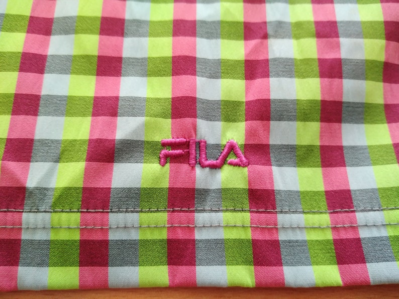 May include: A close-up of a pink, green, and gray checkered fabric with the word "FILA" embroidered in pink.