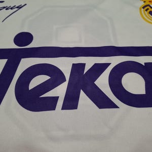 Op de afbeelding: Een wit sportshirt met het paarse Teka-logo prominent op de borst gedrukt. In de rechterbovenhoek bevindt zich het geel-blauwe Real Madrid-embleem en in de linkerbovenhoek staat een zwarte handgeschreven handtekening met de tekst Taguy.