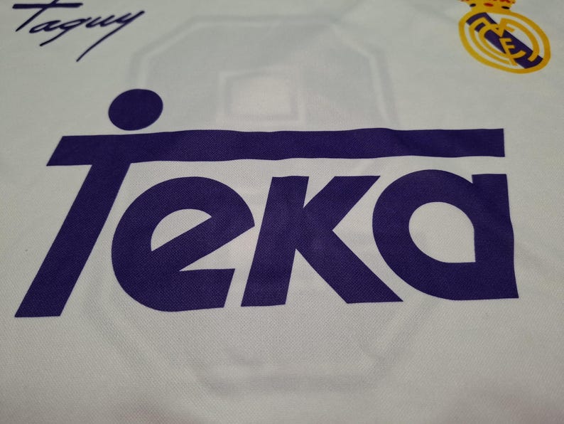 Op de afbeelding: Een wit sportshirt met het paarse Teka-logo prominent op de borst gedrukt. In de rechterbovenhoek bevindt zich het geel-blauwe Real Madrid-embleem en in de linkerbovenhoek staat een zwarte handgeschreven handtekening met de tekst Taguy.