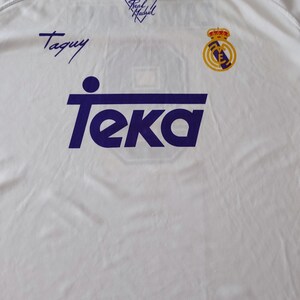 Op de afbeelding: Een wit shirt met korte mouwen en een polokraag met blauwe randen. De voorzijde toont het paarse Teka-logo, het Real Madrid-embleem en de handtekening Taquy. De schouders hebben een blauwe streep met sterren en de tekst Real Madrid.