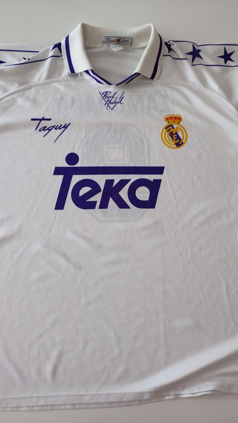 Op de afbeelding: Een wit shirt met korte mouwen en een polokraag met blauwe randen. De voorzijde toont het paarse Teka-logo, het Real Madrid-embleem en de handtekening Taquy. De schouders hebben een blauwe streep met sterren en de tekst Real Madrid.