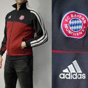 May include: A black and red Adidas track jacket with white stripes on the sleeves. The jacket has a zip fastening and a small FC Bayern Munich logo on the chest.