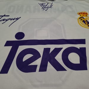 Op de afbeelding: Een wit sportshirt met een groot paars Teka-logo op de voorzijde. Het shirt bevat een Real Madrid-teamembleem in geel en blauw, samen met twee handtekeningen in zwarte inkt, waarvan er &eacute;&eacute;n Taguy leest en de andere Real Madrid.