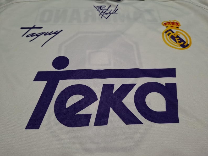 Op de afbeelding: Een wit sportshirt met een groot paars Teka-logo op de voorzijde. Het shirt bevat een Real Madrid-teamembleem in geel en blauw, samen met twee handtekeningen in zwarte inkt, waarvan er &eacute;&eacute;n Taguy leest en de andere Real Madrid.
