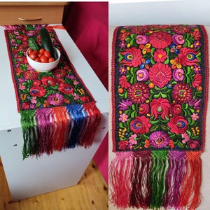 Può includere: Un runner da tavolo nero con fiori ricamati colorati e foglie verdi. Il runner ha una frangia di nappe rosse, rosa, verdi e blu.