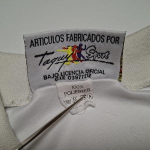 Op de afbeelding: Een witte kledingkraag met een rechthoekig stoffen label met de tekst ARTICULOS FABRICADOS POR Taquy Sport BAJO LICENCIA OFICIAL C.I.F 03977212 en een kleiner label eronder met 100% POLIESTER, wassymbolen en de tekst MADE IN SPAIN.