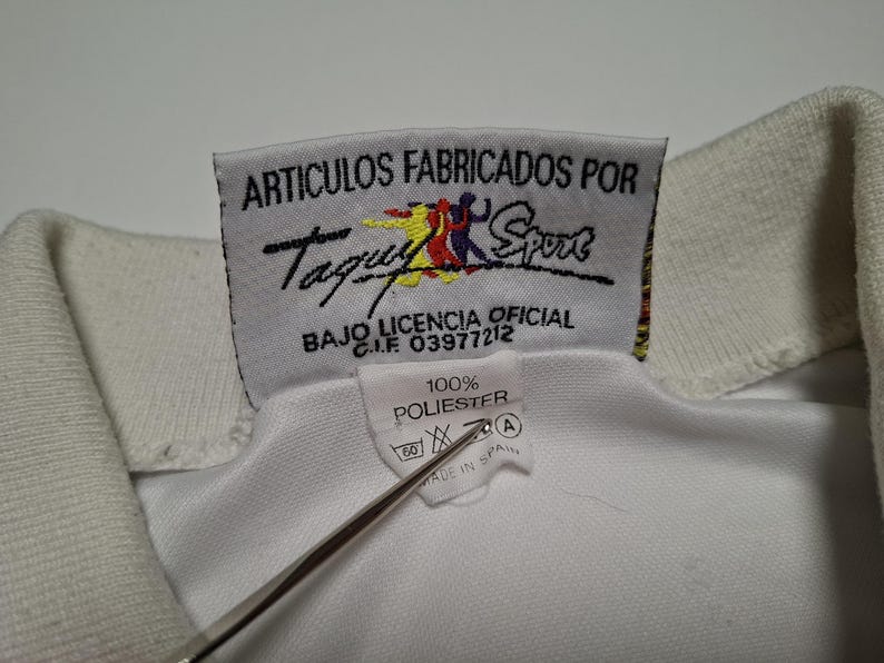 Op de afbeelding: Een witte kledingkraag met een rechthoekig stoffen label met de tekst ARTICULOS FABRICADOS POR Taquy Sport BAJO LICENCIA OFICIAL C.I.F 03977212 en een kleiner label eronder met 100% POLIESTER, wassymbolen en de tekst MADE IN SPAIN.