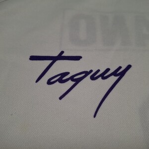 Op de afbeelding: Een wit stoffen oppervlak met een donkerpaarse handtekening die Taguy leest in een cursief handschrift. Het materiaal heeft een zichtbare fijne mesh-textuur en een vaag, gedeeltelijk verborgen woord is op de achtergrond boven de handtekening gedrukt.