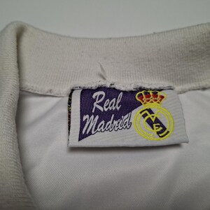 Op de afbeelding: Een witte overhemdkraag met een rechthoekig stoffen label aan de binnenzijde. Het label toont een paarse wimpelvorm met de tekst Real Madrid in wit schrift, vergezeld van een geel en rood kroonembleem en een geel cirkelvormig logo.