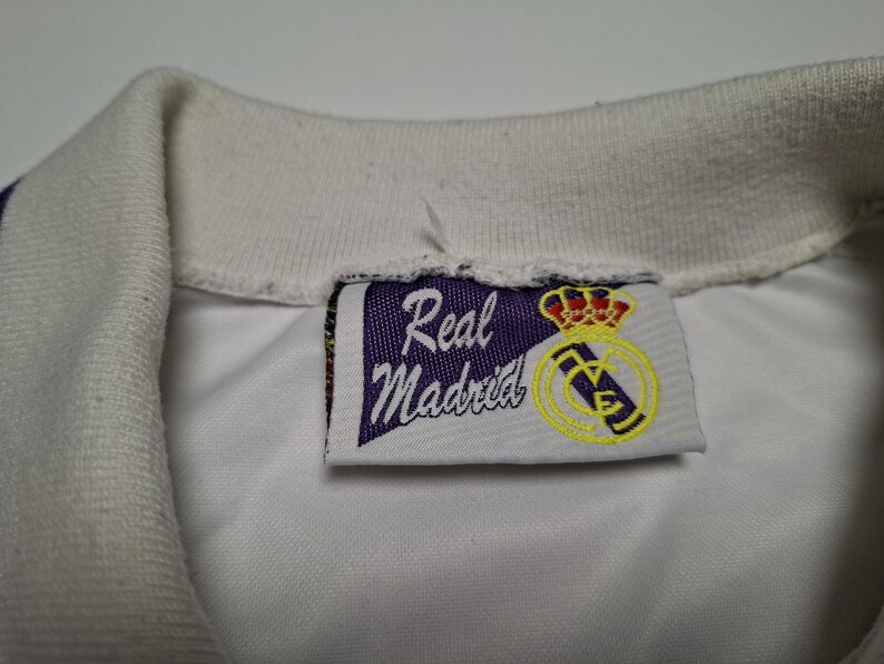 Op de afbeelding: Een witte overhemdkraag met een rechthoekig stoffen label aan de binnenzijde. Het label toont een paarse wimpelvorm met de tekst Real Madrid in wit schrift, vergezeld van een geel en rood kroonembleem en een geel cirkelvormig logo.