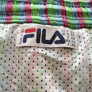 May include: White Fila brand label with red and blue lettering. The label is attached to a pair of white mesh shorts with a green, blue, and pink plaid waistband. The label includes size information: I 50, D 50, F 42, USA 34, J XL.