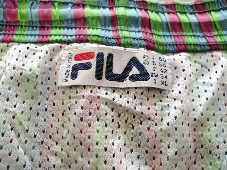 May include: White Fila brand label with red and blue lettering. The label is attached to a pair of white mesh shorts with a green, blue, and pink plaid waistband. The label includes size information: I 50, D 50, F 42, USA 34, J XL.