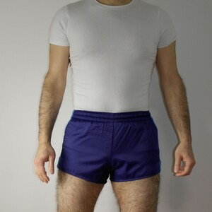 Op de afbeelding: Een man draagt een wit T-shirt en blauwe sport shorts met een witte tailleband en blauwe bies.