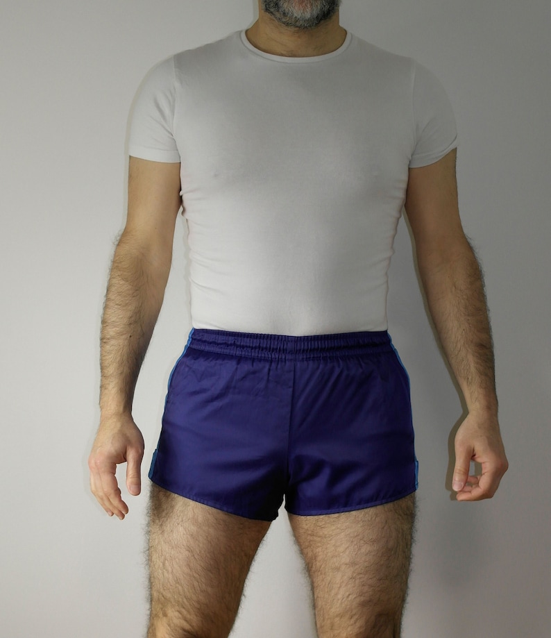 Op de afbeelding: Een man draagt een wit T-shirt en blauwe sport shorts met een witte tailleband en blauwe bies.