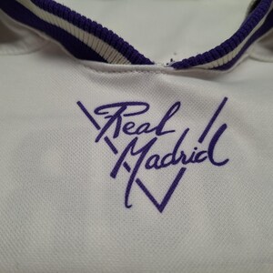 Op de afbeelding: Een wit sportshirt met een paars geborduurd Real Madrid-logo op de borst. De kraag is wit met een paars-witte gestreepte bies. De stof heeft een subtiel textuurpatroon dat over het gehele kledingstuk zichtbaar is.