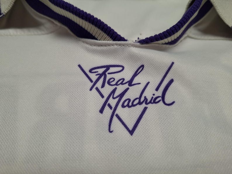 Op de afbeelding: Een wit sportshirt met een paars geborduurd Real Madrid-logo op de borst. De kraag is wit met een paars-witte gestreepte bies. De stof heeft een subtiel textuurpatroon dat over het gehele kledingstuk zichtbaar is.