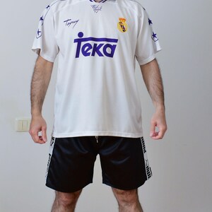 Op de afbeelding: Een wit voetbalshirt met korte mouwen, een paarse kraag, paarse sterren op de schouders, de tekst Teka in het paars en het Real Madrid-embleem. Gecombineerd met een zwarte sportshort met witte zijpanelen en zwarte Puma-branding.
