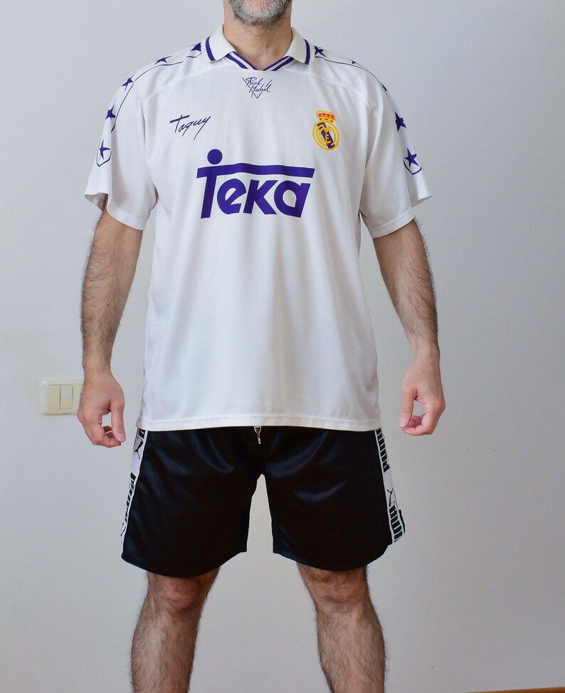 Op de afbeelding: Een wit voetbalshirt met korte mouwen, een paarse kraag, paarse sterren op de schouders, de tekst Teka in het paars en het Real Madrid-embleem. Gecombineerd met een zwarte sportshort met witte zijpanelen en zwarte Puma-branding.