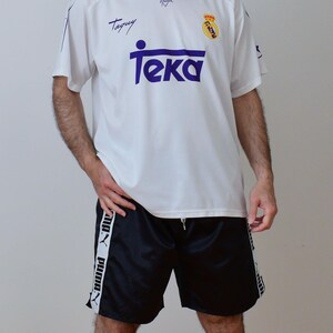 Op de afbeelding: Een wit voetbalshirt met korte mouwen, een paarse kraag, het woord Teka in het paars, een Real Madrid-embleem en de naam Taguy. Gecombineerd met een zwarte sportbroek met een witte verticale streep aan beide kanten met het Puma-logo.
