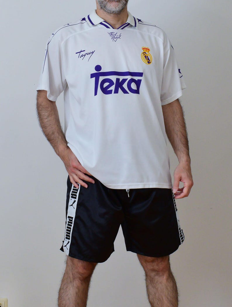 Op de afbeelding: Een wit voetbalshirt met korte mouwen, een paarse kraag, het woord Teka in het paars, een Real Madrid-embleem en de naam Taguy. Gecombineerd met een zwarte sportbroek met een witte verticale streep aan beide kanten met het Puma-logo.