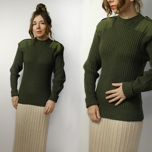 May include: Dark olive green ribbed knit sweater with long sleeves and shoulder patches. The sweater features a crew neck and a fitted shape. The model is wearing a beige skirt. A versatile piece for a casual or vintage-inspired look.