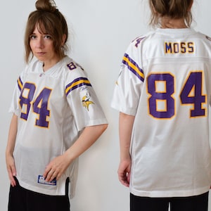 Peut inclure: Un maillot Reebok blanc des Minnesota Vikings avec le numéro 84 en violet et bordure jaune sur le devant et le dos. Le nom MOSS est imprimé en violet au dos. Les manches présentent des rayures violettes et jaunes et un logo des Vikings.