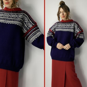 Puede incluir: Un suéter azul marino con un patrón geométrico en blanco y rojo. El suéter tiene cuello redondo y el patrón se repite en las mangas. La modelo lleva pantalones rojos.