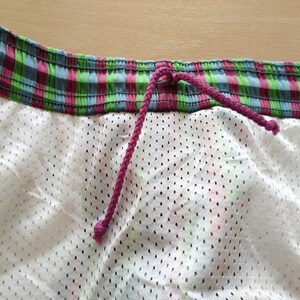 May include: Close-up of the waistband of a pair of swim trunks. The waistband is made of a green, pink, and blue plaid fabric with a pink drawstring.
