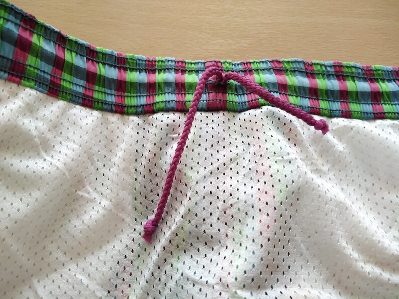 May include: Close-up of the waistband of a pair of swim trunks. The waistband is made of a green, pink, and blue plaid fabric with a pink drawstring.