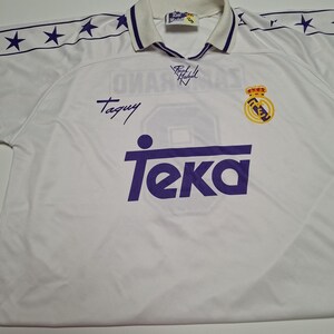 Op de afbeelding: Een wit Real Madrid voetbalshirt met een paars Teka-sponsorlogo op de borst, een teamembleem en een kraag met paarse bies. De schouders hebben een paars sterrenpatroon en de stof bevat handtekeningen en een Real Madrid-label in de nek.