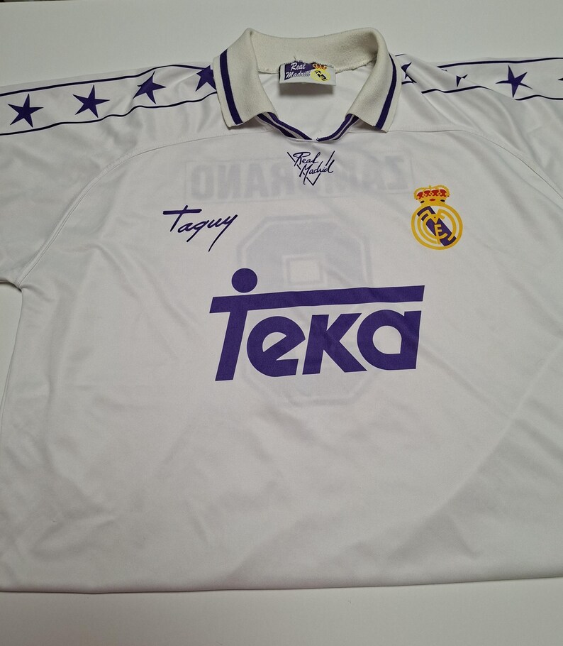 Op de afbeelding: Een wit Real Madrid voetbalshirt met een paars Teka-sponsorlogo op de borst, een teamembleem en een kraag met paarse bies. De schouders hebben een paars sterrenpatroon en de stof bevat handtekeningen en een Real Madrid-label in de nek.