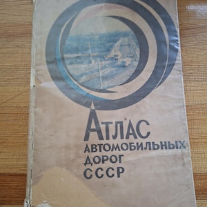 May include: Vintage road atlas with a cover featuring a highway scene within a circular design. The title "ATЛАС АВТОМОБИЛЬНЫХ ДОРОГ СССР" is printed in bold, dark letters. The cover has a worn, aged appearance.