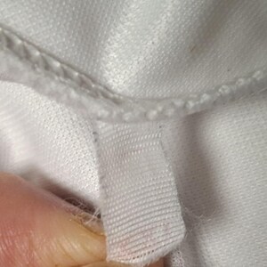 Op de afbeelding: Een close-up van een wit kledingstuk met een gestikte naad en een klein, rechthoekig wit stoffen label aan de binnenrand. Het materiaal heeft een subtiele, fijngebreide textuur met zichtbare draadlussen langs de zoomlijn.