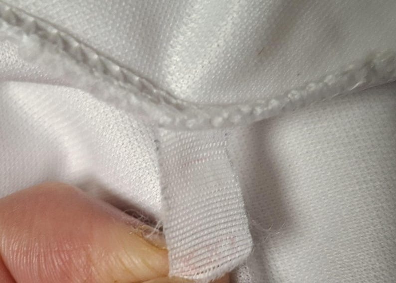 Op de afbeelding: Een close-up van een wit kledingstuk met een gestikte naad en een klein, rechthoekig wit stoffen label aan de binnenrand. Het materiaal heeft een subtiele, fijngebreide textuur met zichtbare draadlussen langs de zoomlijn.