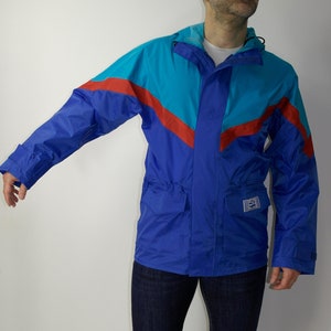 Könnte beinhalten: Eine blaue, rote und türkisfarbene Jacke mit einem Reißverschluss und einer Kapuze. Die Jacke hat eine aufgesetzte Tasche auf der linken Seite und einen Kordelzug in der Taille.
