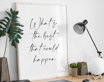 May include: A framed print with the text "What's the best that could happen" in a cursive font. The print is on a wooden desk with a laptop, camera, speaker, lamp, and decorative items. The desk is against a white wall.